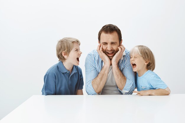 Filmación en interiores de un padre molesto y llorando sentado a la mesa, gritando y con las manos apoyadas en la cara, sintiéndose deprimido e infeliz mientras los hijos hacen ruido y le gritan