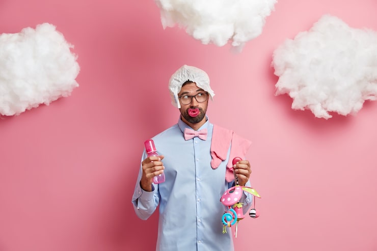 Foto gratuito filmación en interiores de pensativo futuro padre barbudo posa con artículos para bebés lleva camisa de gafas y pañal en la cabeza piensa en el nombre del niño recién nacido
