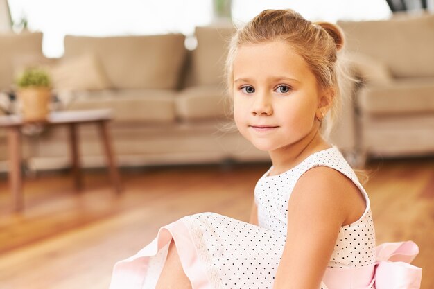 Filmación en interiores de la pequeña princesa linda con un hermoso vestido rosa sentado en el piso en casa preparándose para el desempeño de los niños en el jardín de infantes con adorables ojos azules