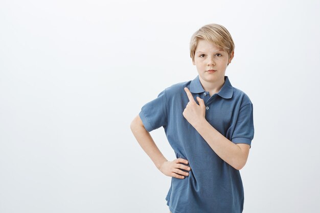 Filmación en interiores de serio lindo niño con cabello rubio en camiseta azul, sosteniendo la mano en la cadera y apuntando a la esquina superior izquierda con el dedo índice