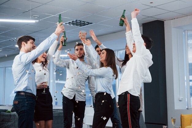 Finalmente lo conseguimos. Foto del equipo joven en ropa clásica que celebra el éxito mientras sostiene bebidas en la moderna oficina bien iluminada