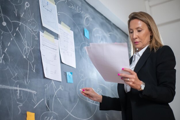 Una financiera seria que analiza el crecimiento empresarial. Vista de ángulo bajo de la mujer analizando datos financieros, dibujando gráficos de crecimiento de ganancias con tiza a bordo. Análisis financiero, concepto de crecimiento empresarial