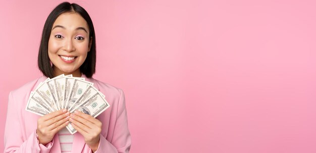 Finanzas microcrédito y concepto de personas Feliz empresaria asiática sonriente mostrando dólares dinero de pie en traje contra fondo rosa