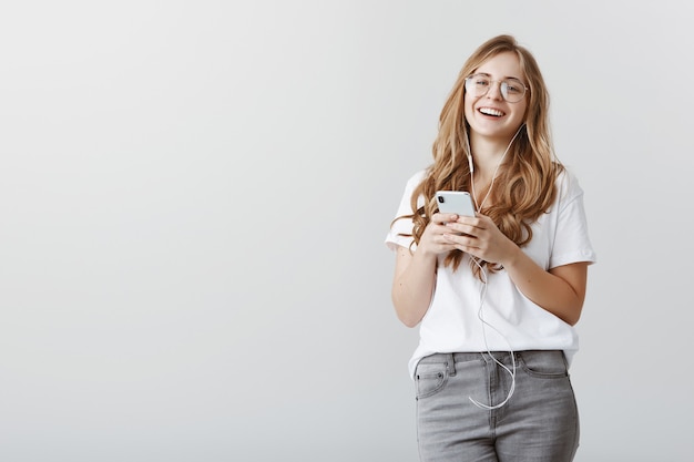 Fingir que te escucha mientras escuchas música. Retrato de mujer europea con estilo atractivo con cabello rubio y gafas, sonriendo positivamente, sosteniendo el teléfono inteligente y usando auriculares sobre la pared gris