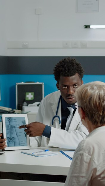 Fisioterapeuta joven que muestra la tableta de la lesión de los huesos a un paciente mayor con osteoporosis en el escritorio en el consultorio del médico. Anciana mirando vértebras virtuales para examen médico