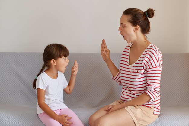 Fisioterapeuta profesional que trabaja en defectos o dificultades del habla con una niña pequeña en el interior mientras está sentada en el sofá, un niño pequeño tiene una lección de idioma para mejorar el habla.