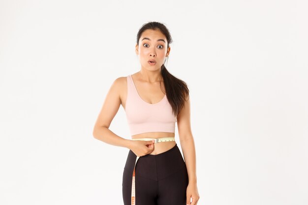 Fitness, estilo de vida saludable y concepto de bienestar. Chica asiática sorprendida en dieta, deportista envuelve cinta métrica alrededor de la cintura y parece impresionada al perder peso con el entrenamiento.