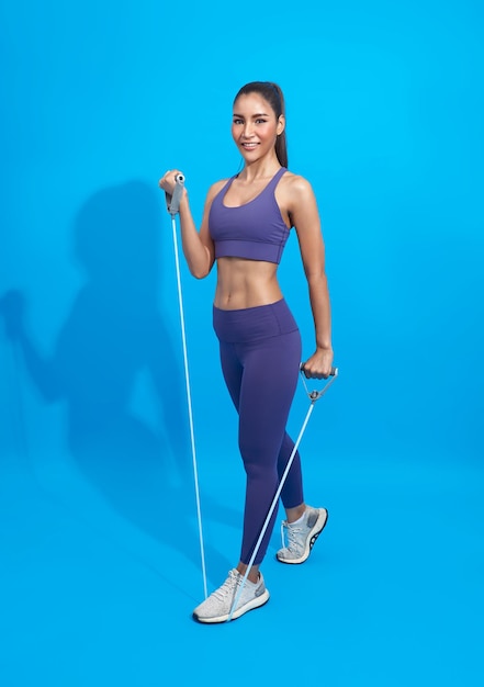 Fitness mujer asiática en ropa deportiva haciendo ejercicio con una banda de resistencia sobre fondo azul.