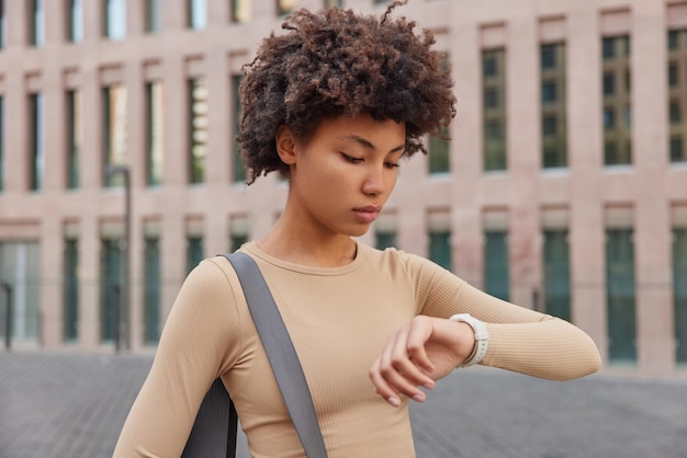 Fitness mujer comprueba los resultados en el reloj inteligente vestido con ropa deportiva beige que lleva karemat enrollado va a hacer deporte regularmente posa cerca de un edificio urbano al aire libre Mujer atlética después de hacer ejercicio