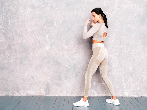 Fitness mujer confiada en ropa deportiva Sexy joven hermosa modelo se encuentra en un estante Mujer posando cerca de la pared gris en el estudio Practica boxeo de golpes de poder durante el proceso de entrenamiento