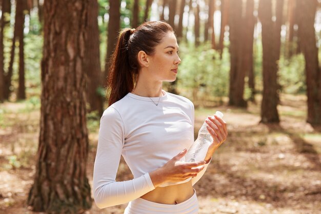 Fitness mujer hermosa con cabello oscuro y cola de caballo sosteniendo una botella de agua y mirando a otro lado, posando después de hacer ejercicio en el bosque