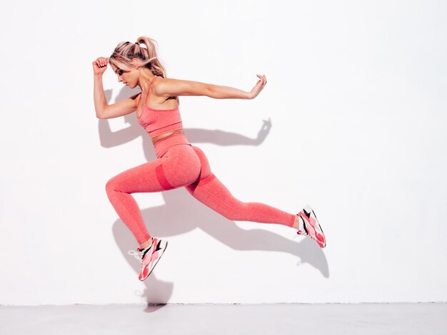 Fitness mujer sonriente en ropa deportiva rosa Joven hermosa modelo con cuerpo perfecto Mujer posando en la calle cerca de la pared blanca en el día soleado de verano Alegre y feliz Saltando y corriendo