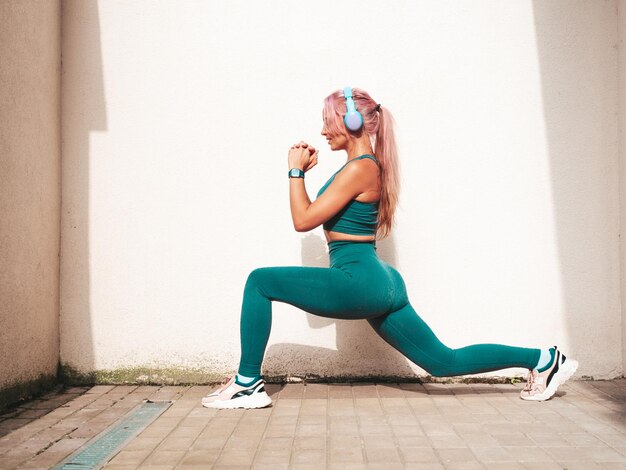 Fitness mujer sonriente en ropa deportiva verde con cabello rosado Joven hermosa modelo con cuerpo perfecto Mujer posando en la calle cerca de la pared blanca Escuchando música en auriculares inalámbricos