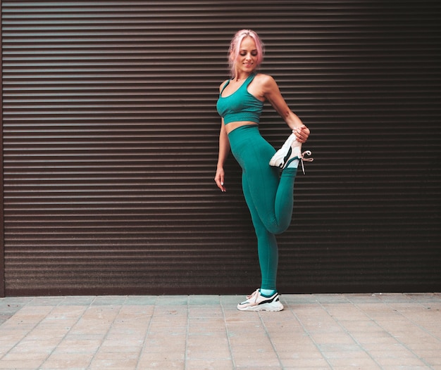 Fitness mujer sonriente en ropa deportiva verde con cabello rosado Modelo hermosa joven con cuerpo perfecto Mujer posando en la calle cerca de la pared de persiana Estirándose antes de entrenar