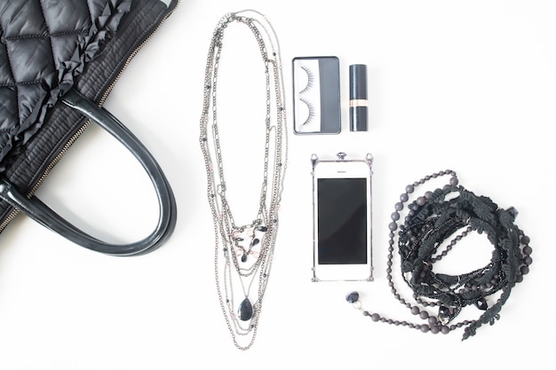 Flat lay de cuaderno vacío, teléfono móvil y accesorios de mujer en concepto de negro, vista superior, aislado sobre fondo blanco