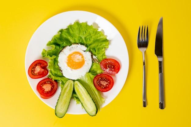 Flat lay fried huevo con plato de verduras frescas con cubiertos sobre fondo liso