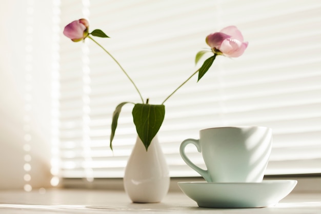 Flor de jarra y taza de café con platillo cerca de la persiana de la ventana