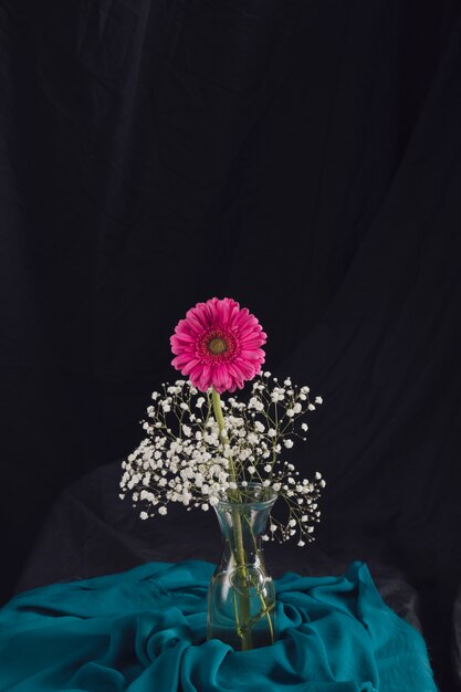 Flor rosa con ramitas de flores en un jarrón cerca de un textil azul en la oscuridad
