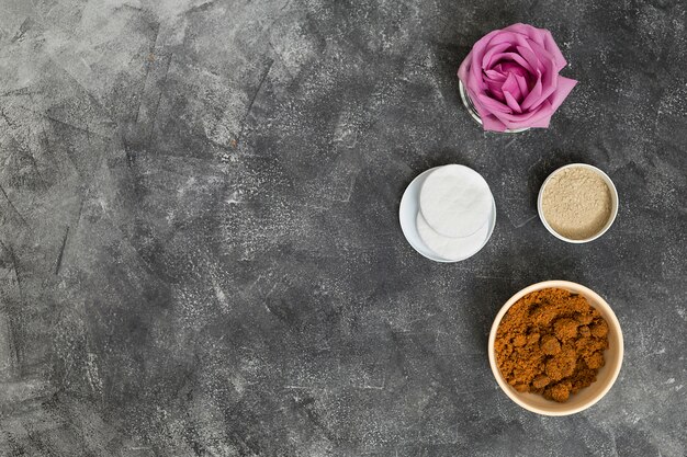 Flor rosa rosa; Almohadillas de algodón y tazones de café en polvo y arcilla rhassoul sobre fondo de hormigón gris