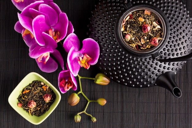 Flor rosada de la orquídea y hierba seca del té con la tetera de cerámica de la textura en el placemat negro