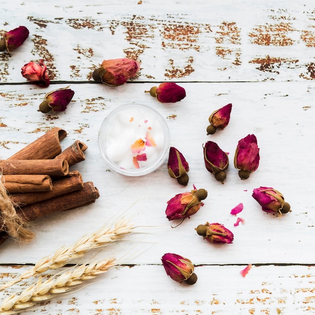 Flor de te de capullos de rosa; palitos de canela; algodón en un tazón; gavilla de espigas de trigo en tablón de madera de textura blanca