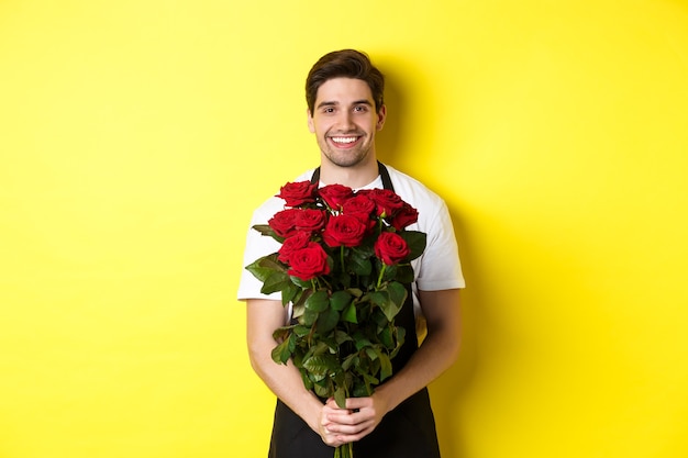 Floreria sonriente en delantal negro sosteniendo flores vendiendo ramo de rosas sobre fondo amarillo