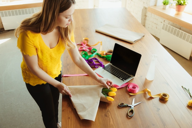 Floreria en el trabajo: mujer muestra cómo hacer un ramo de tulipanes. Joven mujer caucásica da un taller en línea de hacer regalos, presentes para la celebración. Trabajar en casa mientras está aislado, concepto en cuarentena.