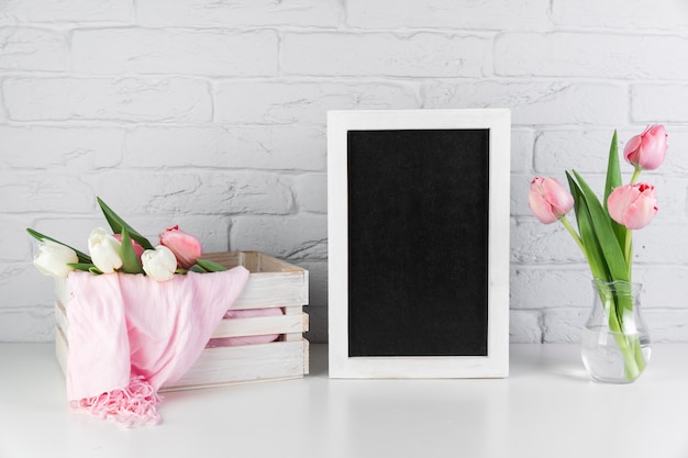 Florero y caja de tulipanes cerca del marco de borde blanco negro en blanco en el escritorio contra la pared de ladrillo