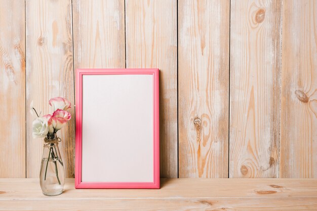 Florero cerca del marco blanco en blanco con borde rosado contra la pared de madera