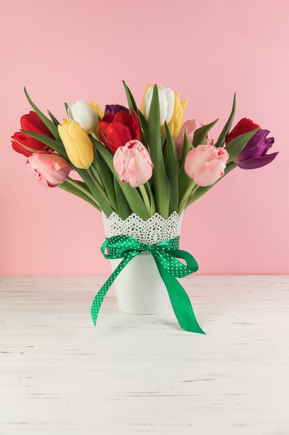 Florero colorido de los tulipanes con el arco verde en el escritorio de madera contra fondo rosado