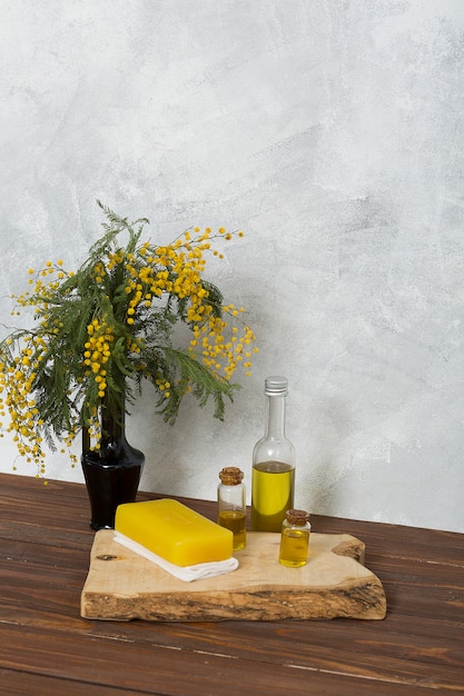 Florero de mimosa amarillo con jabón herbal y una botella de aceite esencial en una tabla de madera sobre una mesa contra la pared gris