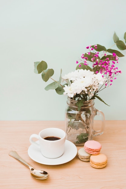 Florero; taza de cafe; Cuchara y macarrones en mesa de madera con fondo de color