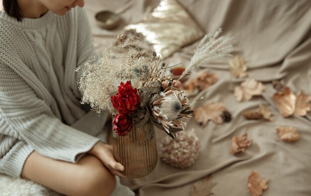 Florero de vidrio con flores de otoño en manos femeninas sobre un fondo borroso con hojas de otoño.