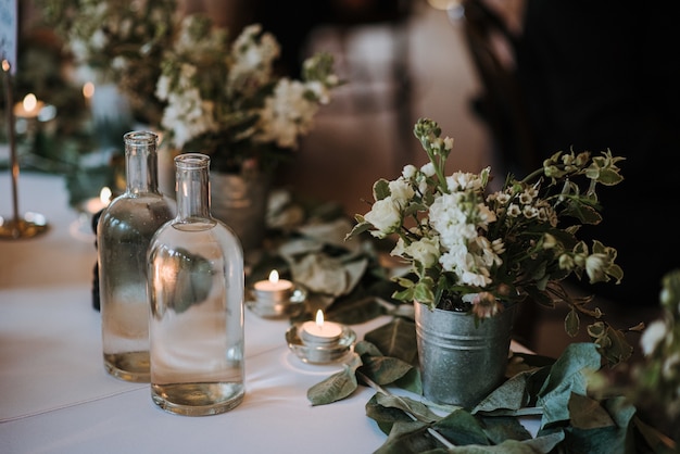 Flores blancas en un balde, botellas de agua y velas en una mesa decorada con hojas