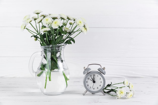Flores blancas de crisantemo en frasco de vidrio cerca del pequeño reloj despertador en el escritorio de madera