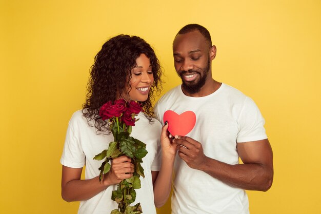 Flores y corazón. Celebración del día de San Valentín, feliz pareja afroamericana aislada sobre fondo amarillo de estudio. Concepto de emociones humanas, expresión facial, amor, relaciones, vacaciones románticas.