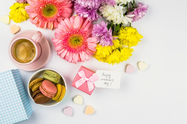 Flores frescas cerca de la etiqueta con palabras en presente, taza de bebida y macarrones en un tazón