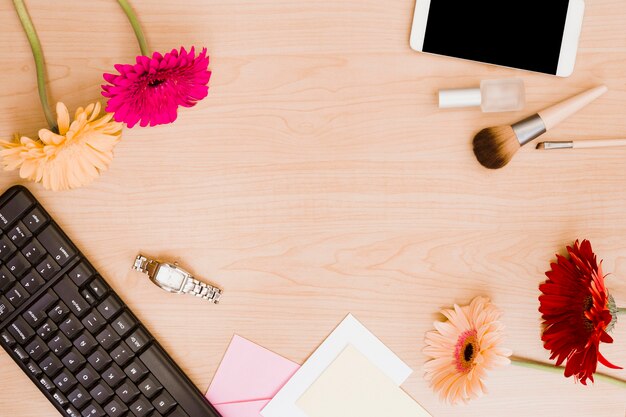 Flores de gerbera; teclado; reloj de pulsera; sobre; brocha de maquillaje; botella de esmalte de uñas y teléfono móvil en el escritorio de madera