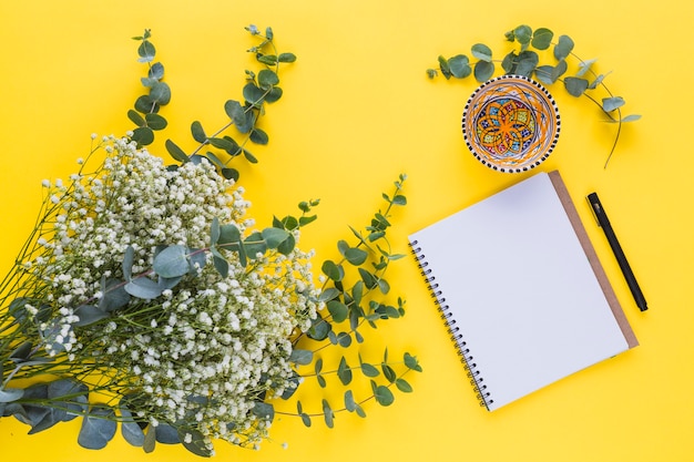 Flores de gypsophila con hojas; bloc de notas espiral cuenco; pluma sobre fondo amarillo