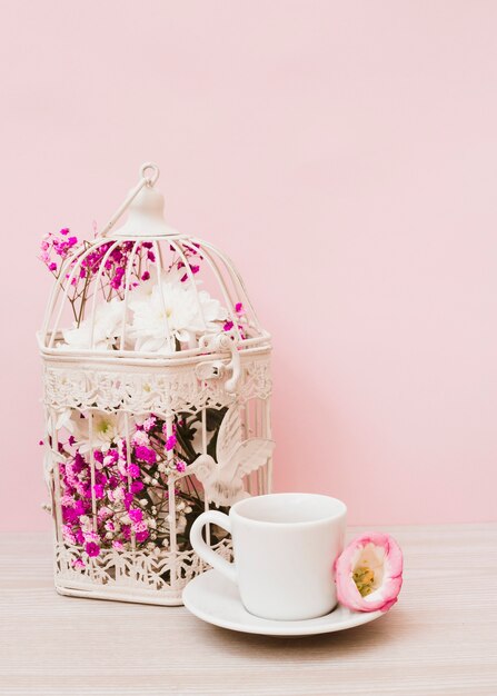 Flores hermosas en la jaula blanca con la taza y el platillo en el escritorio de madera contra fondo rosado