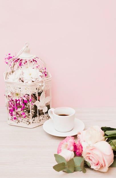 Flores en jaula blanca; Taza de café y rosas en el escritorio de madera con fondo rosa