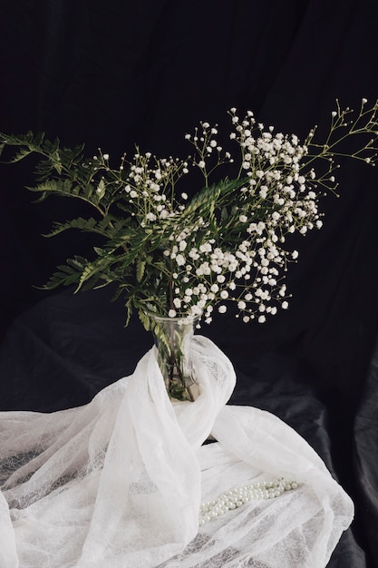 Foto gratuita flores con plantas en jarrón junto a textil blanco y cuentas.