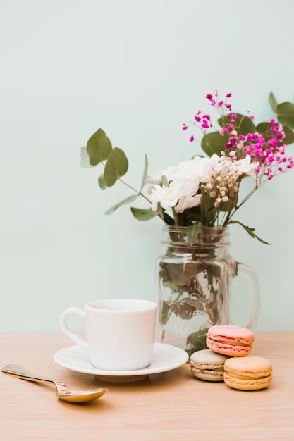 Flores en tarro con taza; Cuchara y macarrones en el escritorio de madera contra la pared.