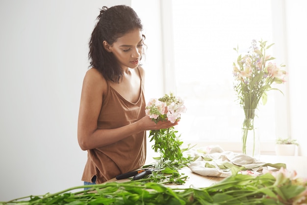 Florista africano atractivo de la mujer que sonríe haciendo el ramo de flores en el lugar de trabajo sobre la pared blanca.