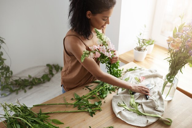 Florista africano atractivo de la mujer que sonríe haciendo el ramo de flores en el lugar de trabajo sobre la pared blanca.