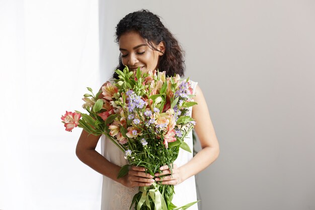 Florista africano tierno de la mujer que sonríe sosteniendo el ramo de alstroemerias sobre la pared blanca. Ojos cerrados.