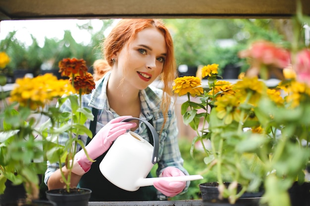 Florista hermosa joven en delantal y guantes rosas de pie con regadera y mirando soñadoramente en cámara mientras trabaja en invernadero