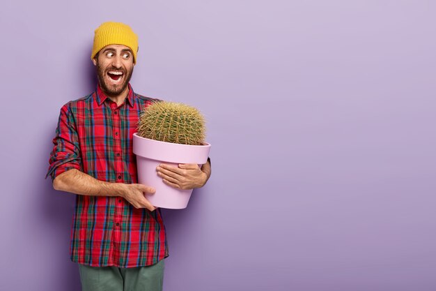 El florista masculino emocional abre la boca ampliamente, sostiene una maceta de cactus espinosos, usa un sombrero amarillo y una camisa a cuadros, posa sobre un fondo púrpura, le gusta cultivar plantas de interior, es muy emocional