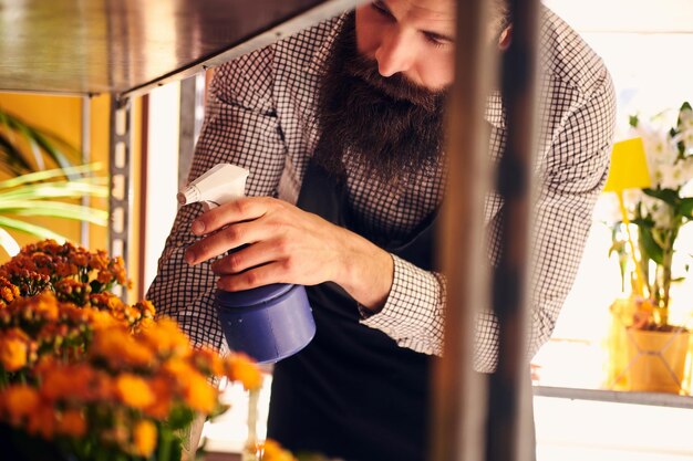 Florista masculino profesional con barba y tatuaje en la mano usando uniforme cuidando flores en una floristería moderna.