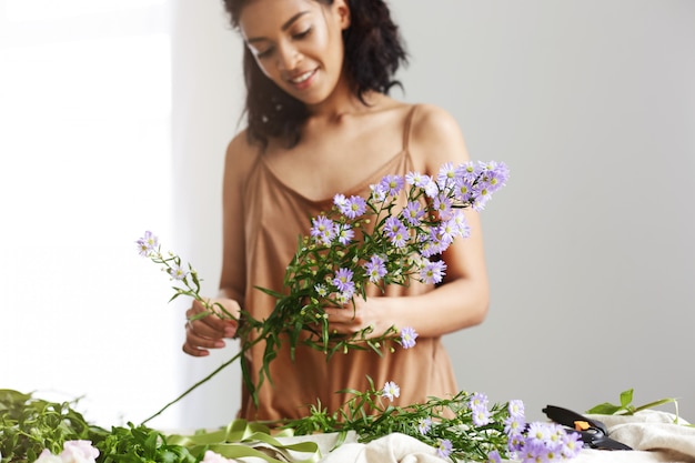 Florista de sexo femenino africano alegre que sonríe trabajando sosteniendo la rama de la margarita sobre la pared blanca.